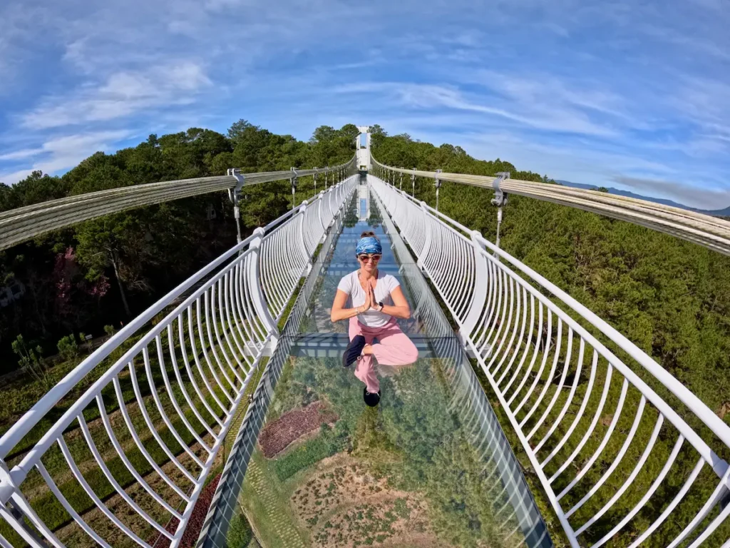 A tourist in a yoga pose on the transparent Ngàn Thông Glass Bridge in Dalat, Vietnam, suspended high above a pine forest under a blue sky. The view is wide-angle, showing the full length of the suspension bridge.