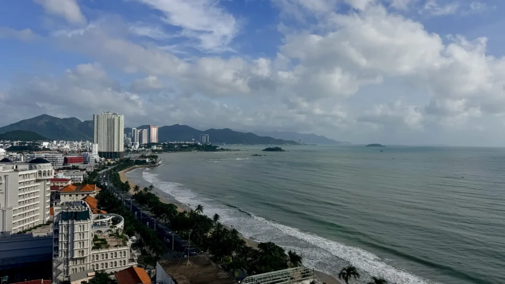 Вид сверху на пляж Нячанга, дорогу Чан Фу и высотные отели на фоне гор и облачного неба.
