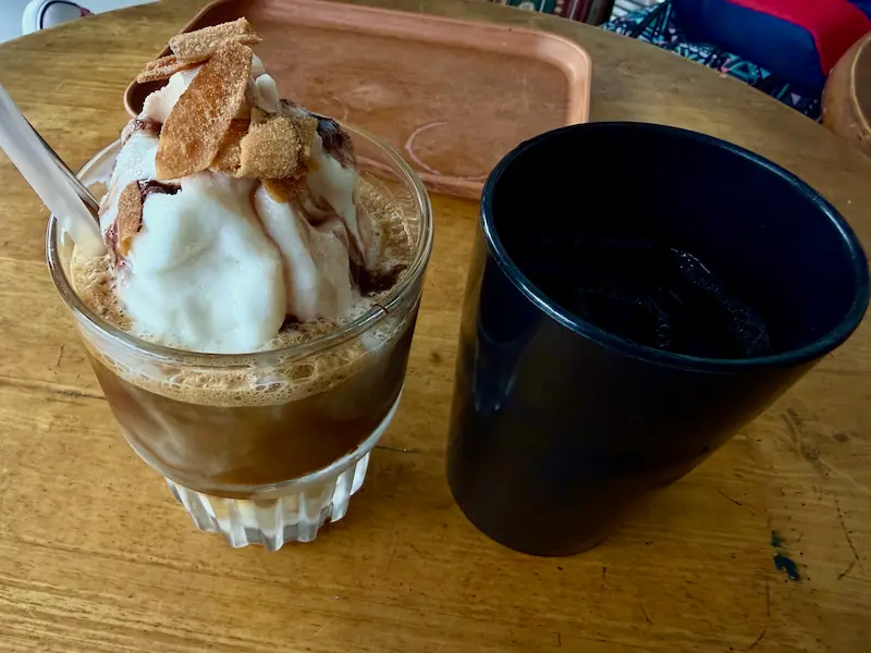 Vietnamese coconut coffee in a glass with toasted coconut toppings served with a side of tea on a wooden cafe table in Nha Trang.