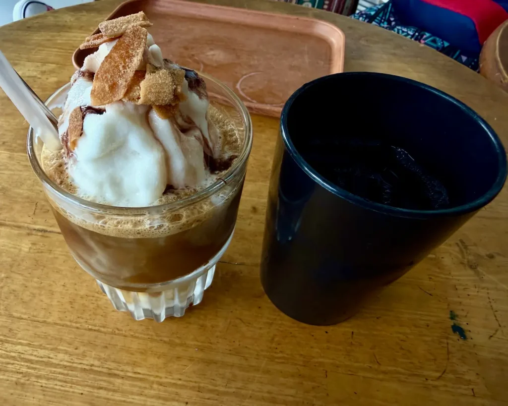 Vietnamese coconut coffee in a glass with toasted coconut toppings served with a side of tea on a wooden cafe table in Nha Trang.