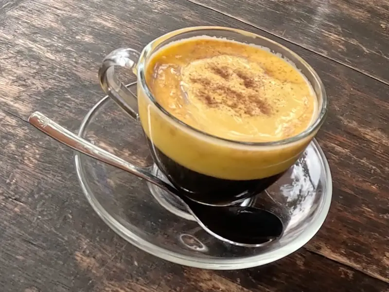 Стеклянная чашка с вьетнамским яичным кофе (Ca Phe Trung) на деревянном столе. Напиток состоит из нижнего слоя темного кофе и густой желтой яичной пенки, посыпанной какао.