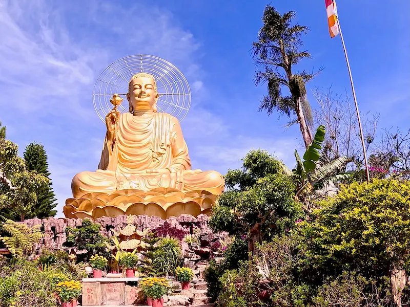 Гигантская статуя сидящего Золотого Будды с цветком лотоса в окружении пышного тропического сада на фоне синего неба в Далате, Вьетнам