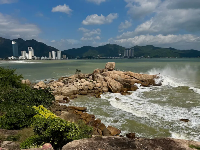 Живописный вид на Сад камней Хон Чонг в Нячанге с гигантскими валунами, разбивающимися волнами и панорамой города с небоскребами на фоне гор.