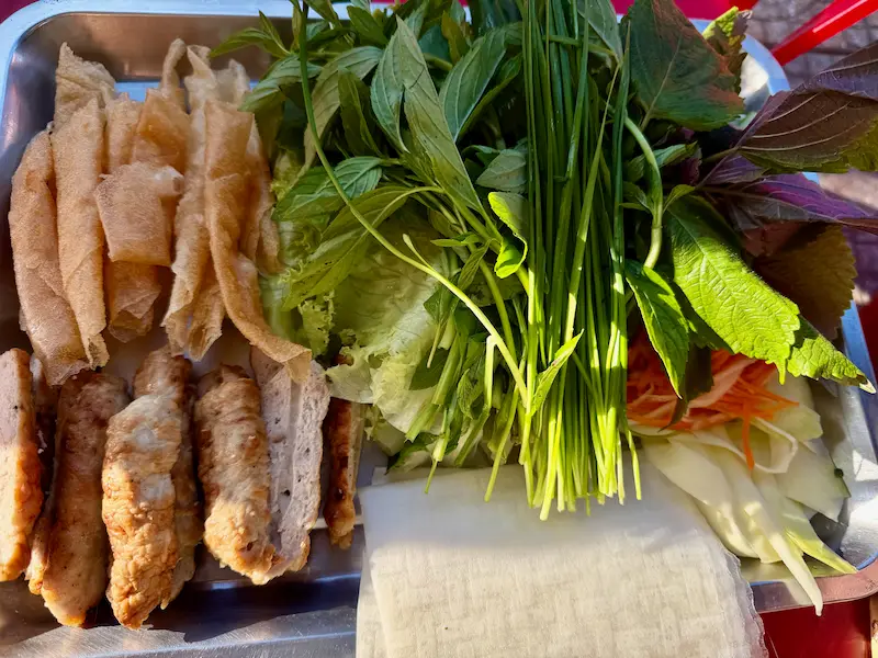 Металлический поднос с ингредиентами для вьетнамского блюда Нэм Нуонг в Нячанге: жареная свинина на гриле, хрустящие трубочки из теста, свежая зелень, маринованная морковь и стопка рисовой бумаги.