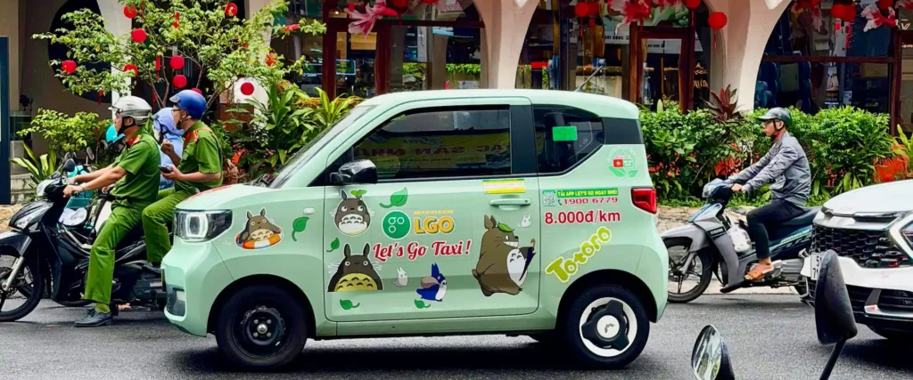 Компактное зеленое электрическое такси LGO с наклейками Тоторо едет по оживленной улице среди мопедов в Нячанге, Вьетнам