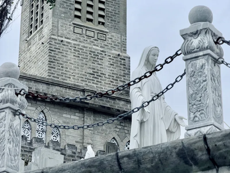 Белая статуя Девы Марии на фоне серой каменной колокольни Кафедрального собора в Нячанге.