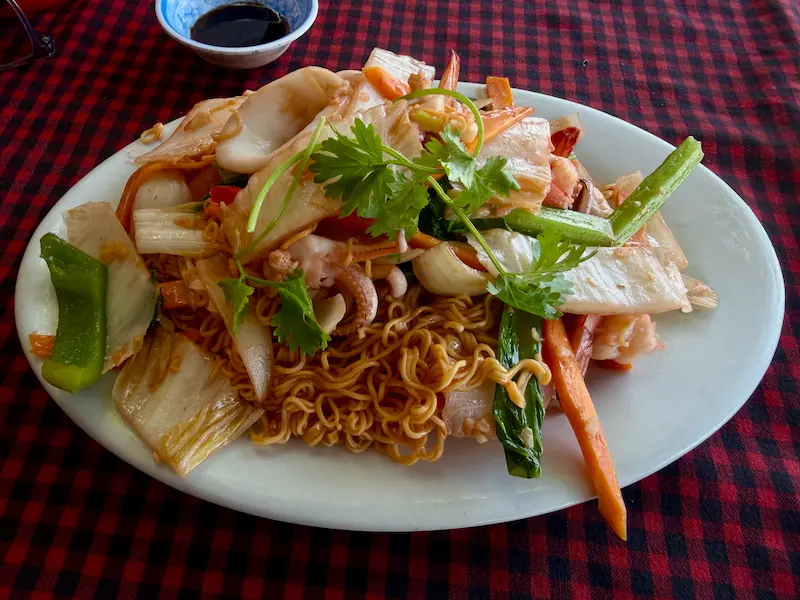 Вьетнамская жареная лапша с морепродуктами (Mì Xào Hải Sản) на белой тарелке. Блюдо щедро украшено крупными кусками белого кальмара, креветками, морковью, капустой бок-чой и свежей кинзой в местном кафе Нячанга.