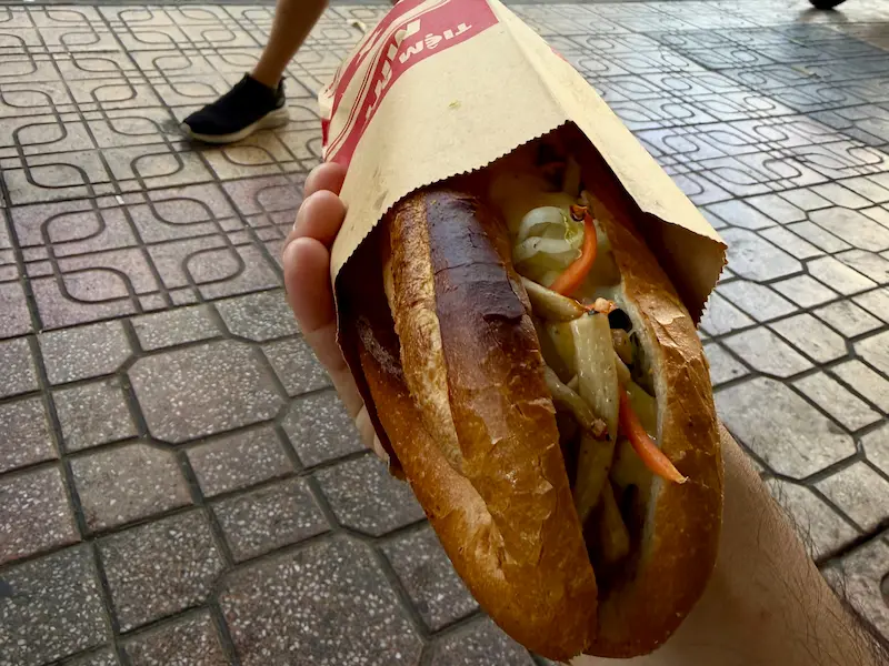 Рука держит свежий вьетнамский сэндвич Бань Ми (Bánh Mì) в бумажном пакете на улице Нячанга. Внутри поджаристого хрустящего багета видна начинка из овощей и грибов.