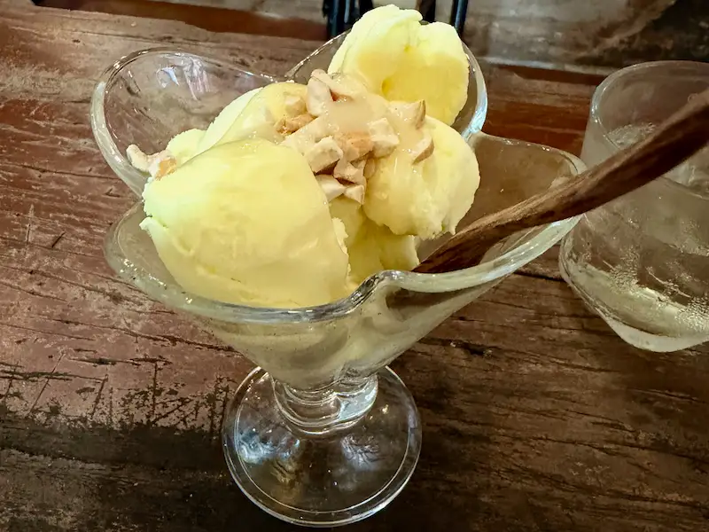 Светло-желтые шарики мороженого с дурианом (Kem Sầu Riêng) в стеклянной креманке на фактурном деревянном столе. Десерт полит сгущенным молоком, посыпан арахисом и подается с деревянной ложечкой в кафе Нячанга.
