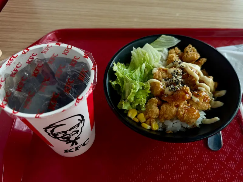 Набор KFC в Нячанге: чаша с хрустящим куриным попкорном, рисом, листьями салата и кукурузой, политая соусами и посыпанная кунжутом. Рядом стоит запечатанный стакан газировки Pepsi (Пепси) со льдом. Все это подано на типичном для фастфуда красном пластиковом подносе.