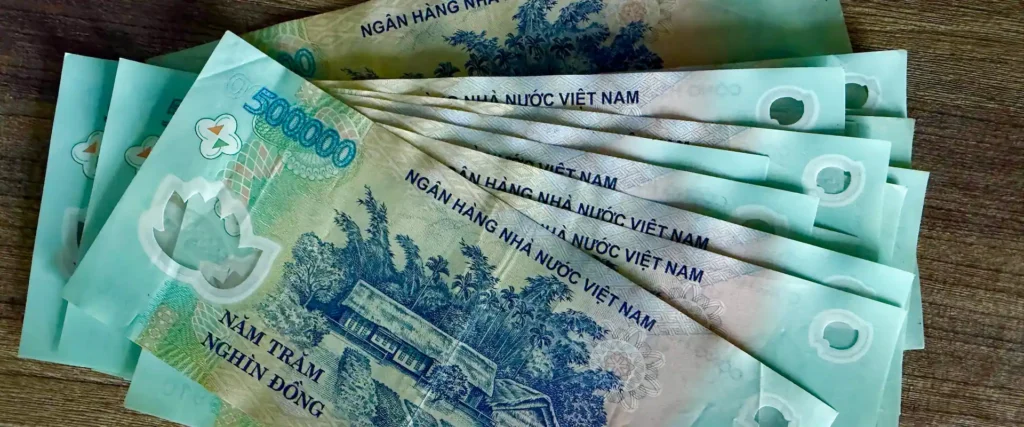 Пачка вьетнамских донгов (VND) на столе, в основном банкноты номиналом 500 000 донгов. На лицевой стороне видны официальные надписи «NGÂN HÀNG NHÀ NƯỚC VIỆT NAM» и «NĂM TRĂM NGHÌN ĐỒNG». Это самая крупная банкнота во Вьетнаме.