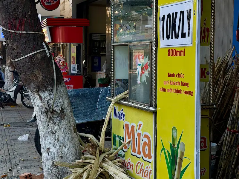 Ярко-желтая уличная тележка для продажи освежающего тростникового сока (Nước Mía) в Нячанге. Четкая вывеска с ценой 10,000 за чашку объемом 800 мл. Видны стебли тростника рядом с деревом.