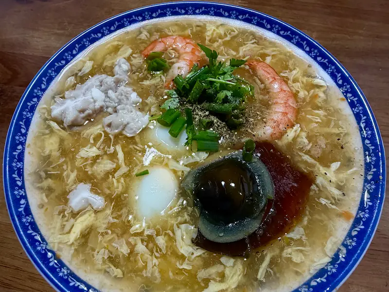 Вид сверху на глубокую сине-белую миску густого вьетнамского крабового супа "Суп Куа" (Súp Cua). В супе видны крупные креветки, перепелиные яйца, столетнее яйцо, зеленый лук, кинза и черный перец.