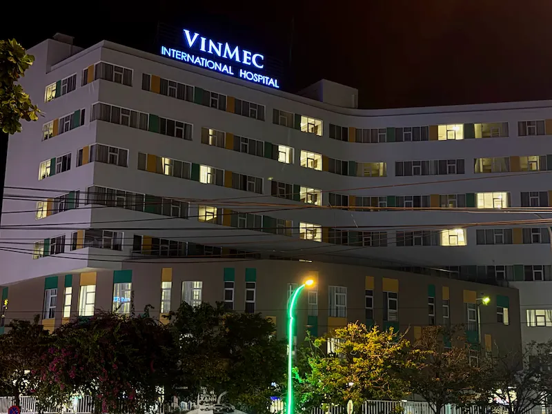 Ночной фасад современного многоэтажного здания клиники Vinmec International Hospital со светящейся синей вывеской в Нячанге, Вьетнам