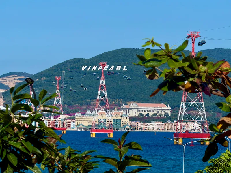 Красно-белые опоры знаменитой канатной дороги над морем, ведущей к парку развлечений VinWonders Nha Trang на фоне гор с белой надписью VINPEARL в Нячанге, Вьетнам